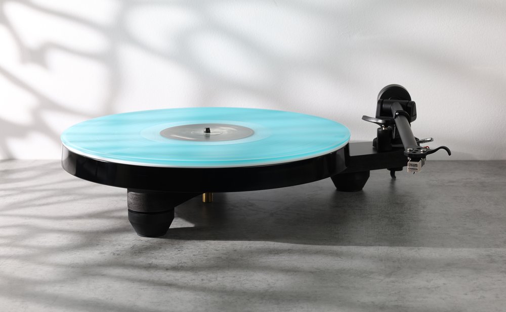 The Rega Replay in matt black at home on a stone surface, a turquoise record on the platter, leaf shadows on the wall behind.