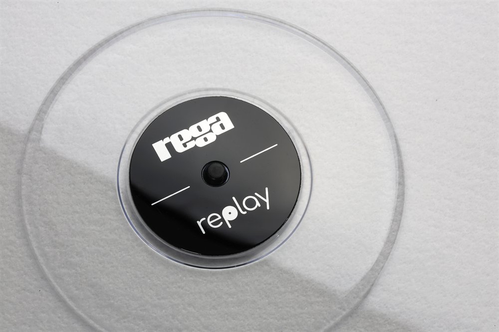 Détail de l'emblème de la Replay : le disque central noir aux logos Rega et Replay, dans de l'acrylique transparent au-dessus d'un tapis blanc.